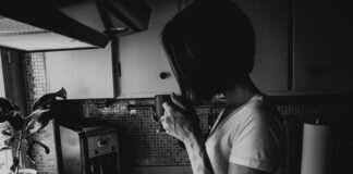 Black and white image of woman whose face cannot be seen, sipping from a mug in a kitchen; her hand is veiny and she seems tense, for Survivor Lit, a literary magazine for sexual assault survivors.